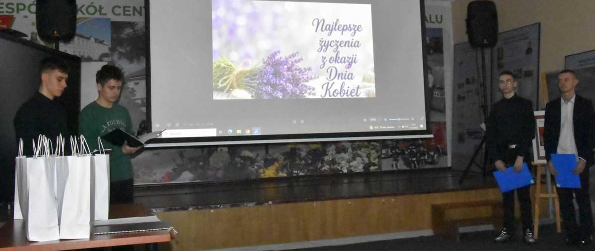 Na zdjęciu widać cztery osoby stojące obok siebie w pomieszczeniu. Trzy z nich trzymają w rękach białe torby upominkowe. W tle znajduje się ekran projekcyjny z wyświetlonym obrazem przedstawiającym gałązkę lawendy oraz napis w języku polskim: „Najlepsze życzenia z okazji Dnia Kobiet”. Po bokach ekranu widoczne są fragmenty plakatów lub banerów z napisami. Scena wygląda na moment wręczania upominków podczas szkolnej lub lokalnej uroczystości.