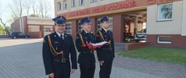 Na zdjęciu znajduje się trzech strażaków w umundurowaniu reprezentacyjnym. Poczet flagowy ustawiony frontem do masztu flagowego. Na rękach jednego z funkcjonariuszy złożona flaga państwowa. 