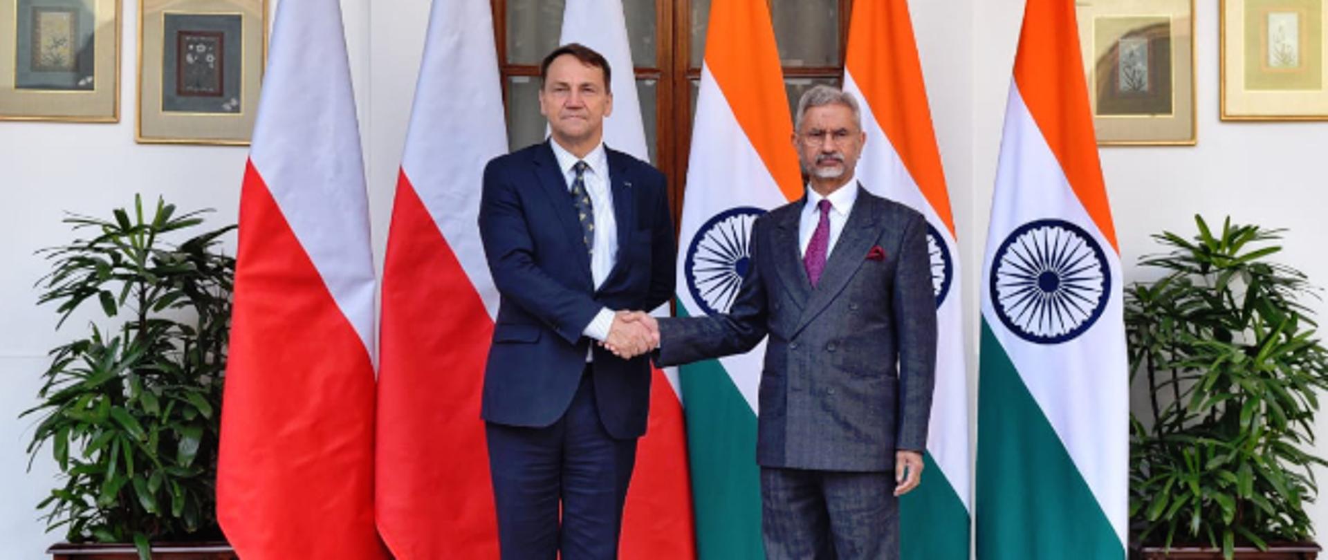 Dwaj mężczyźni w garniturach stoją naprzeciw siebie i podają sobie ręce podczas oficjalnego spotkania. Za nimi widoczne są flagi Polski i Indii, ustawione w reprezentacyjnym wnętrzu.