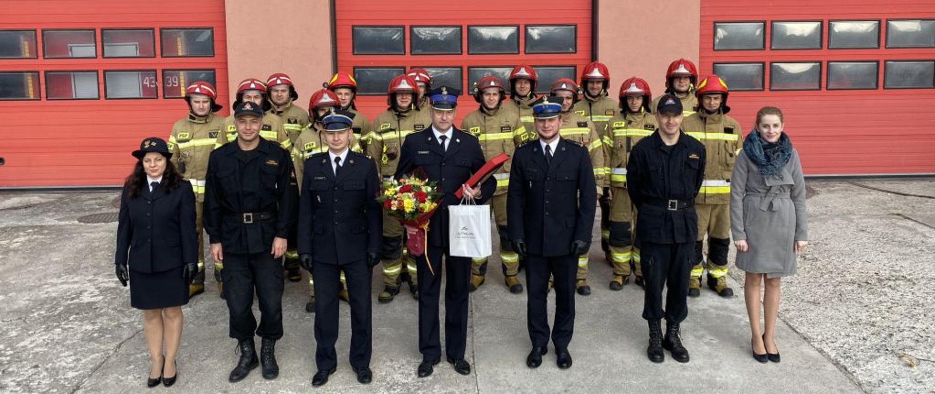 Na tle czerwonych trzech bram garażowych stoją strażacy w dwuszeregu w żółtych ubraniach bojowych. Przed nimi w rzędzie stoją strażacy w granatowych mundurach. Strażak stojący po środku trzyma upominki i kwiaty.