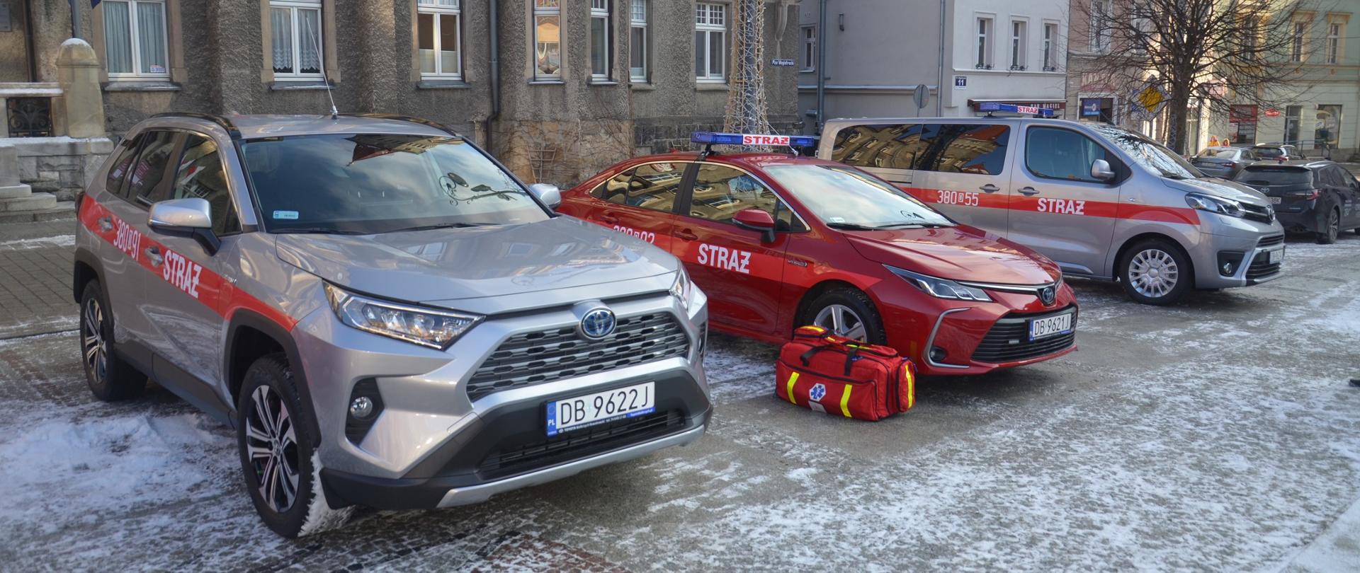 Dwa srebrne i jeden czerwony samochód straży pożarnej stoją na placu. W tle ratusz Urzędu Miasta Wałbrzych