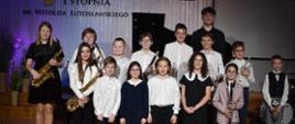 Na zdjęciu widnieje grupa uczniów, ustawionych w dwóch rzędach na scenie auli PSM I stopnia w Jaśle. Niektórzy z uczniów trzymają w rękach saksofony, klarnety i trąbki. Przeważająca kolorystyka zdjęcia: brązowo-czarno-biała. 