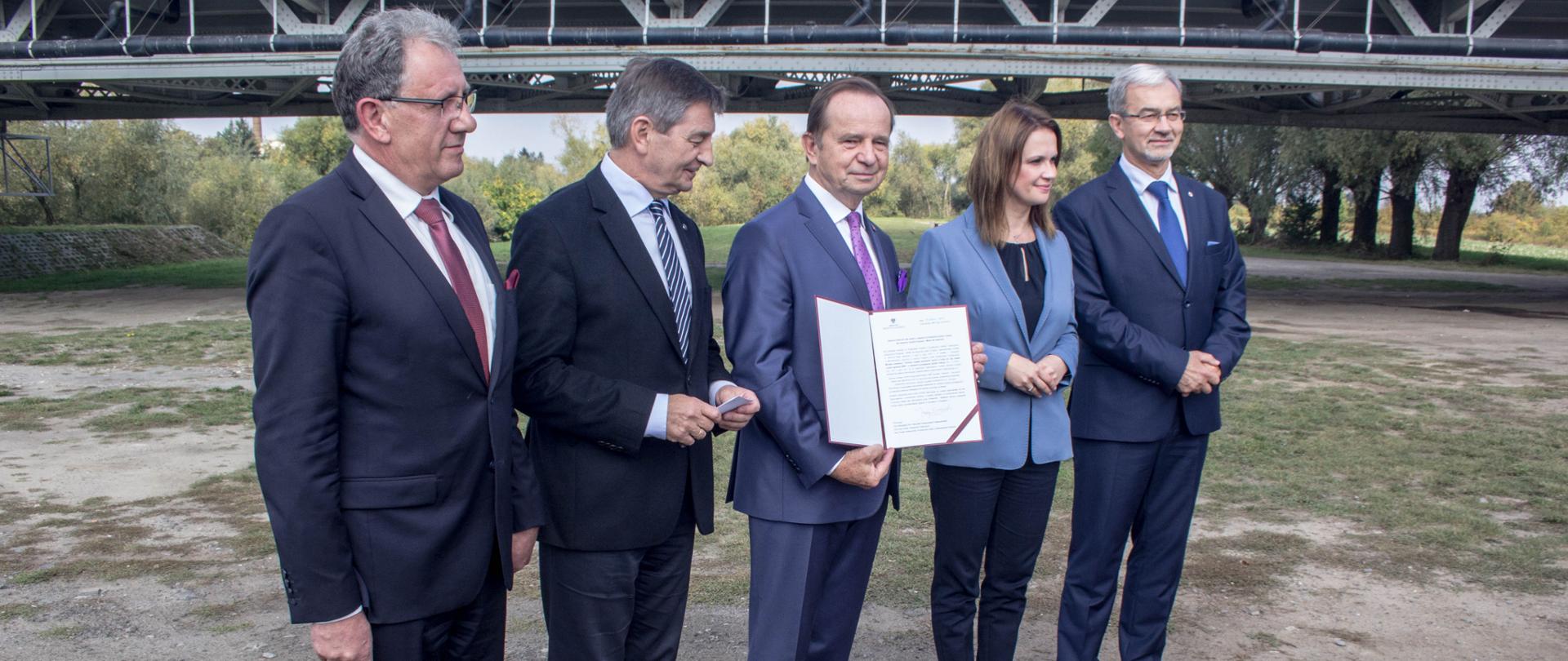 Minister Kwieciński z pozostałymi uczestnikami spotkania stoi, starosta w ręku trzyma podpisaną umowę, w tle widać stary most