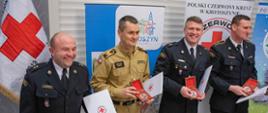 Zdjęcie przedstawiajace czterech strażaków trzymających w rękach medal okolicznościowy. Medale zostały przyznane za dotychczasową działalność na rzecz Polskiego Czerwonego Krzyża. Strażacy w mundurach wyjściowych stoją na tle baneru PCK