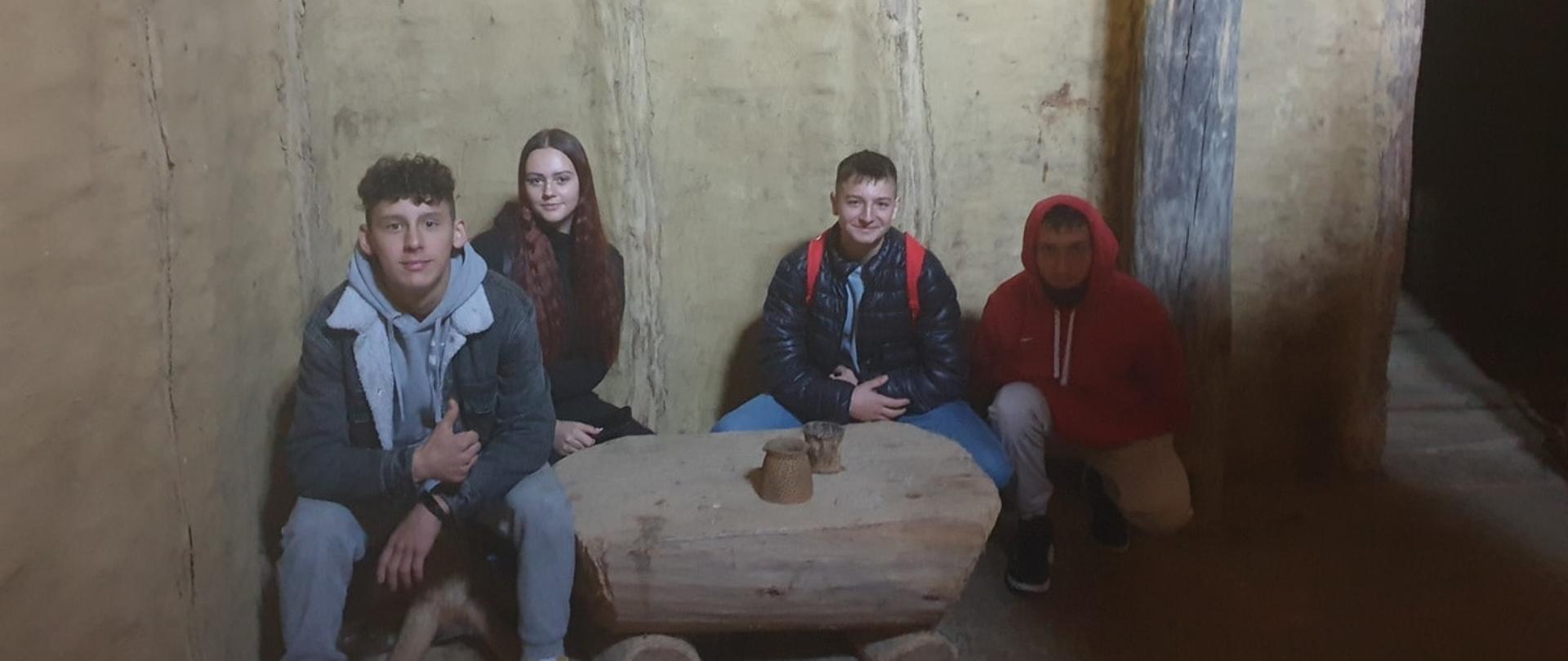 Czworo nastolatków siedzi wewnątrz surowego pomieszczenia o glinianych ścianach. Przed nimi znajduje się prosty, drewniany stół wykonany z pnia drzewa.