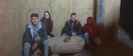 Czworo nastolatków siedzi wewnątrz surowego pomieszczenia o glinianych ścianach. Przed nimi znajduje się prosty, drewniany stół wykonany z pnia drzewa.