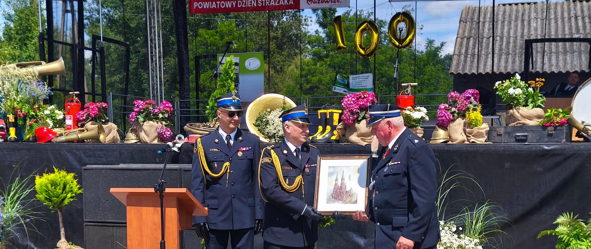 Zastępca Mazowieckiego Komendanta Wojewódzkiego PSP wręcza pamiątkowy obraz druhowi OSP. Obok nich inny strażak w mundurze galowym i drewniana mównica z mikrofonem. Wszyscy stoją na tle dużej sceny z kwiatami i dekoracjami. 