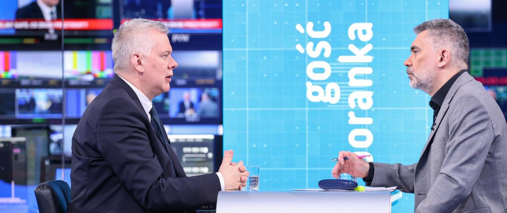 Przy stole siedzą dwie osoby - po lewej minister Tomasz Siemoniak, po prawej - redaktor Mariusz Piekarski. Na stole szklanka z wodą i materiały. W tle widać pionowy napis "Gość Poranka" oraz monitory.