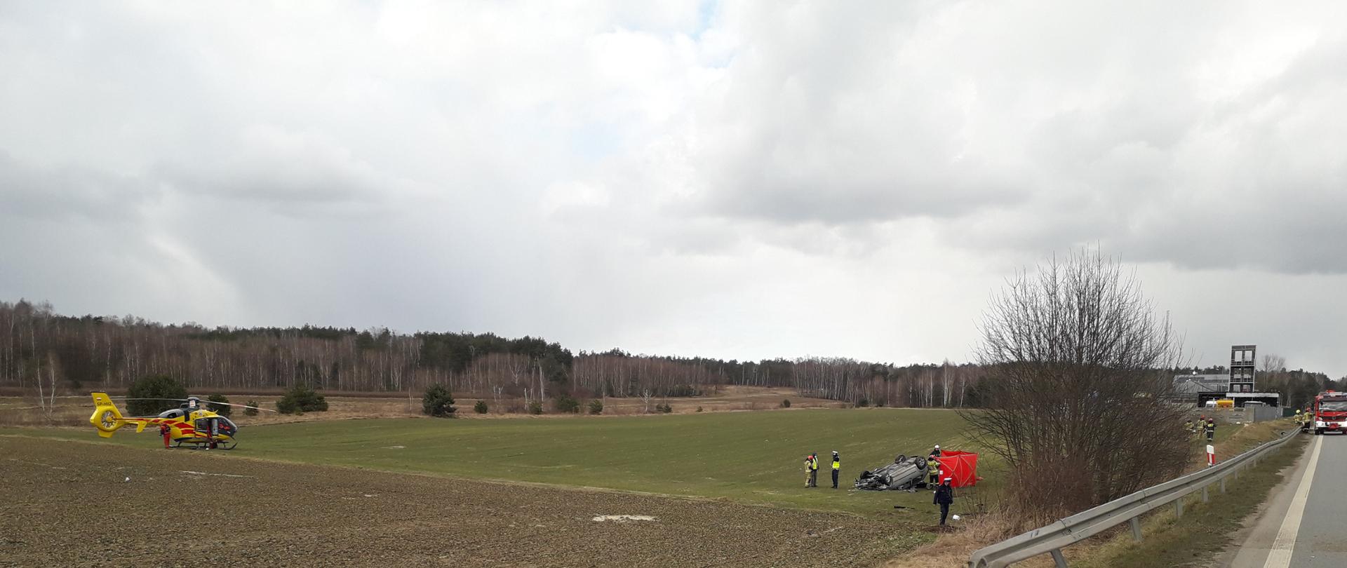 Na zdjęciu w głębi widoczny uszkodzony samochód osobowy, w pozycji na dachu. Wokół pojazdu strażacy oraz policjanci prowadzą czynności ratownicze, strażacy rozstawiają parawan ochronny do osłony miejsc wypadków. W lewej części zdjęcia śmigłowiec Lotniczego Pogotowia Ratunkowego.