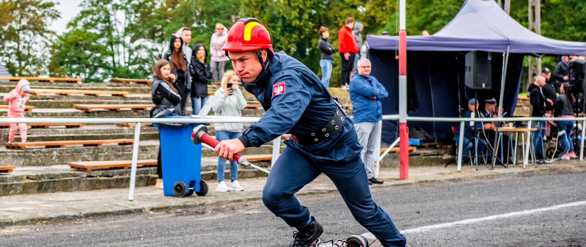 stadion sportowy bieżnia strażak odbiega od przeszkody na bieżni ustawiony jest rozdzielacz oraz wąż w tle inne osoby obserwujące.