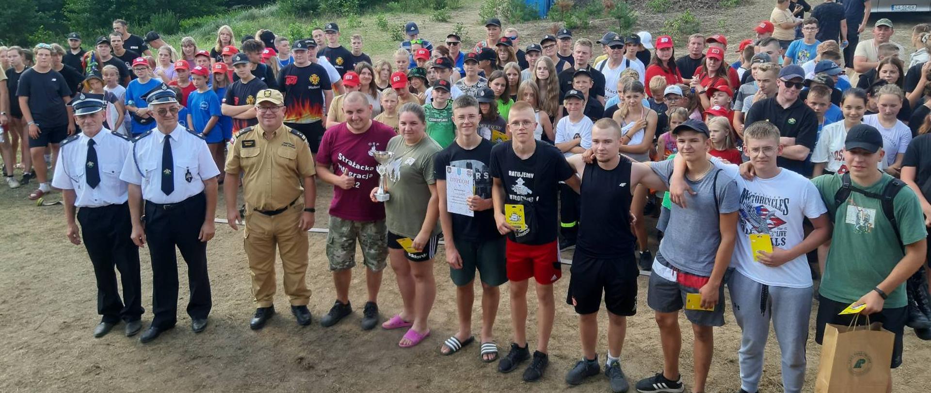 Zdjęcie przedstawia zaproszonych gości i nagrodzonych uczestników. W tle inni uczestnicy marszu, las, samochody pożarnicze