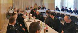 Zdjęcie przedstawia kilkudziesięciu strażaków ubranych w ubrania wyjściowe koloru granatowego, koszule białe, którzy uczestniczą w naradzie operacyjno-szkoleniowej. Siedzą przy stołach . W tle strażak ubrany w umundurowanie służbowe koloru żółtego stoi za stołem i przemawia. W tle widać zasłony w oknach koloru szarego