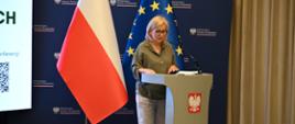 Na zdjęciu Minister Klimatu i Środowiska - Paulina Hennig-Kloska, która stoi za szarą mównicą ozdobioną godłem Polski i patrzy na leżące przed nią dokumenty. Kobieta jest ubrana w luźną koszulę w kolorze khaki, jasne spodnie oraz ciemne buty na obcasie. Po jej prawej stronie stoją dwie duże flagi: polska oraz Unii Europejskiej. W tle widoczna jest ciemnoniebieska ścianka z powtarzającym się logo Ministerstwa Klimatu i Środowiska.