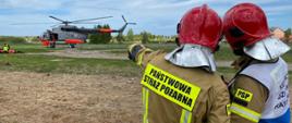 Ćwiczenie RENEGADE/SAREX-21 z udziałem Państwowej Straży Pożarnej - dwóch strażaków PSP stoi tyłem , jeden z nich wskazuje na stojący dalej helikopter
