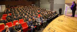 Ujęcie panoramiczne sali audytoryjnej z widokiem na licznie zgromadzonych strażaków w galowych mundurach oraz zaproszonych gości. Na scenie, z prawej strony, widoczny oficer PSP przemawia z mównicy, a w tle, przy ścianie, stoi funkcjonariusz pełniący wartę honorową.
