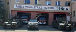 Na tle różowego budynku strażnicy PSP w Hajnówce Z przodu stoją trzy zielone pojazdy wojskowe z tyłu trzy czerwone strażackie wozy bojowe.