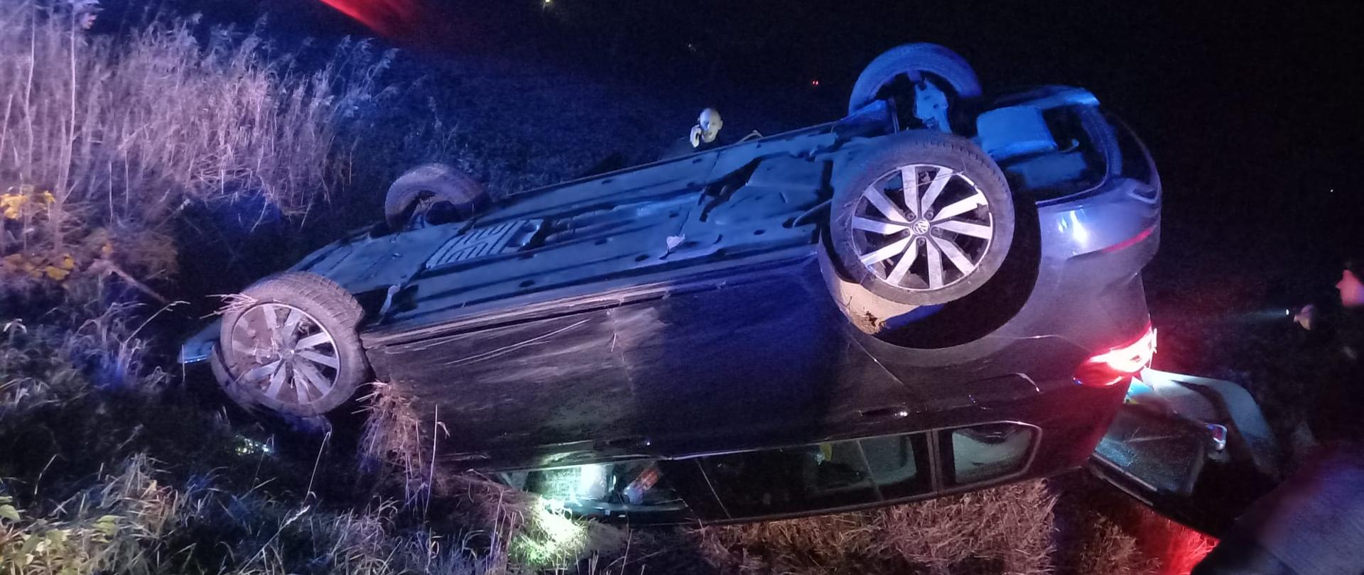 Widać ciemny samochód na dachu, jest w rowie, jest ciemno, miejsce oświetlone przez straż