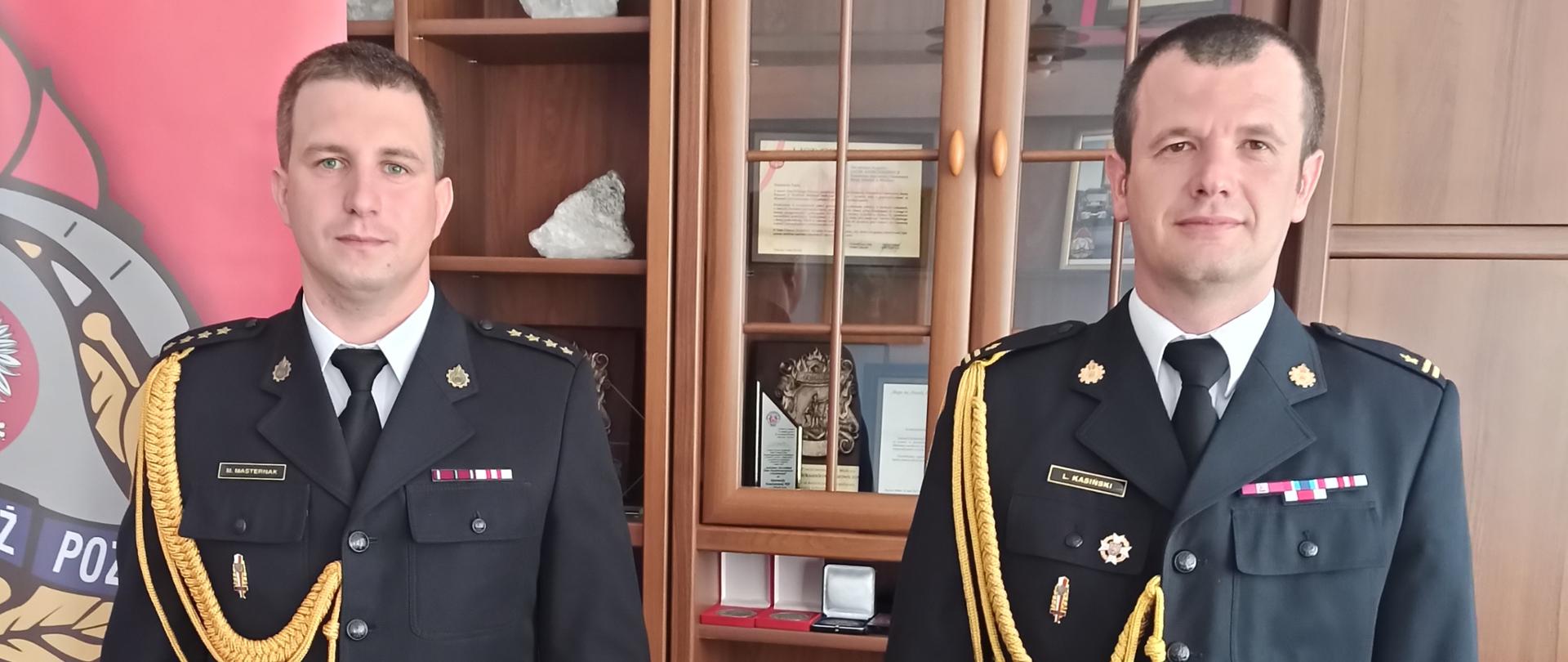 Dwaj strażacy w umundurowaniu wyjściowym. W tle wyposażenie biura.