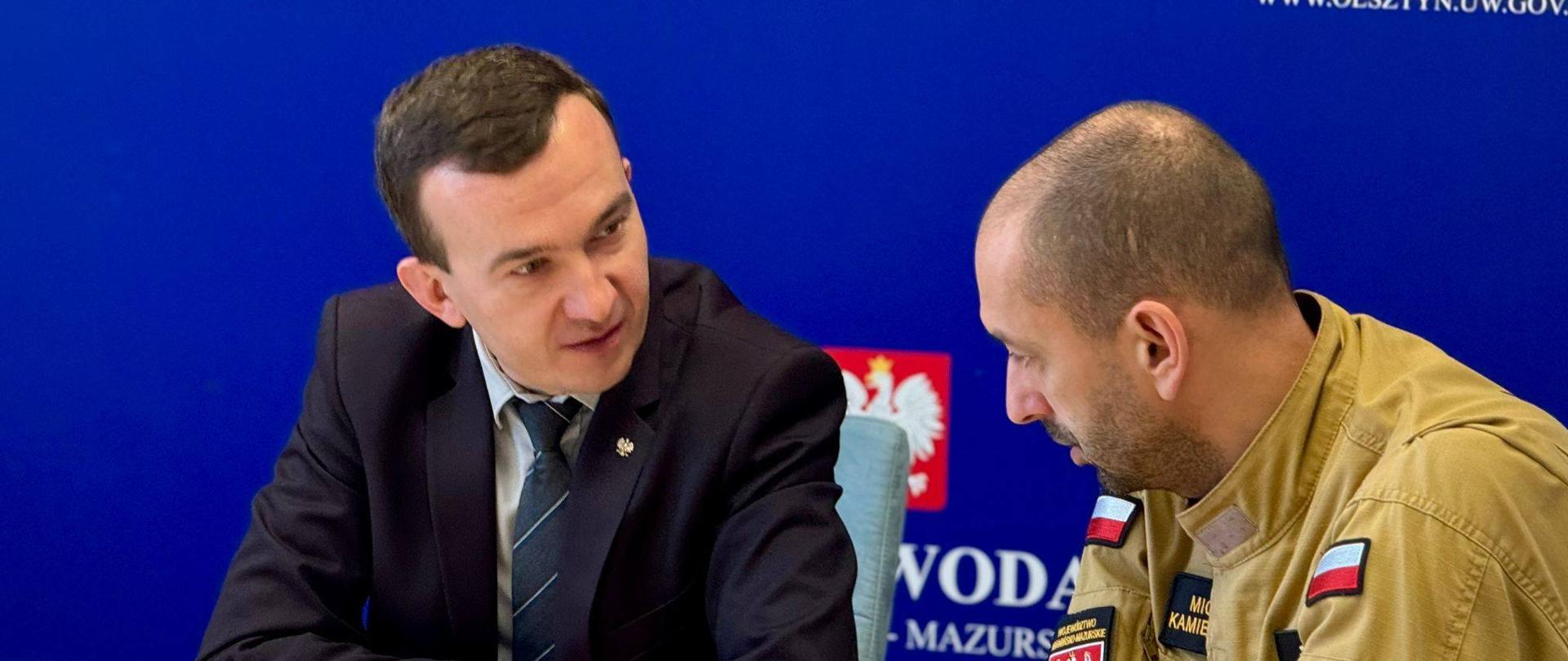 Dwóch mężczyzn, jeden w mundurze strażackim. Rozmawiają.