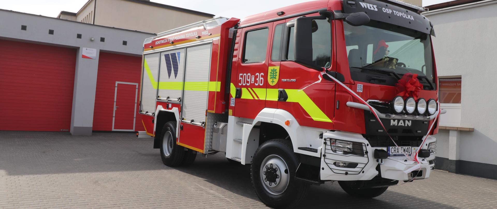 Pojazd pożarniczy, udekorowany biało-czerwonymi wstążkami, wystawiony przed remizę.