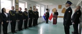 Strażacy PSP w czarnych mundurach wyjściowych oraz pracownicy cywilni stoją wzdłuż dwóch ścian pomieszczenia. Na tle drewnianych dekoracji - Godła Polski, logo ZOSP oraz logo PSP przy mównicy przemawia zastępca Mazowieckiego Komendanta Wojewódzkiego PSP st. bryg. Maciej Bieńczyk. Obok niego stoi Komendant Powiatowy PSP w Lipsku bryg. Artur Kucharski. 