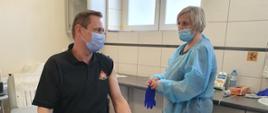 Na zdjęciu strażak ubrana w ubranie koszarowe oraz koszulkę koloru czarnego, siedzi na łóżku w gabinecie lekarskim. Po prawej stronie pielęgniarka ubrana w fartuch podaje szczepionkę w lewe ramię strażaka. Osoby zabezpieczone w maseczki jednorazowe.