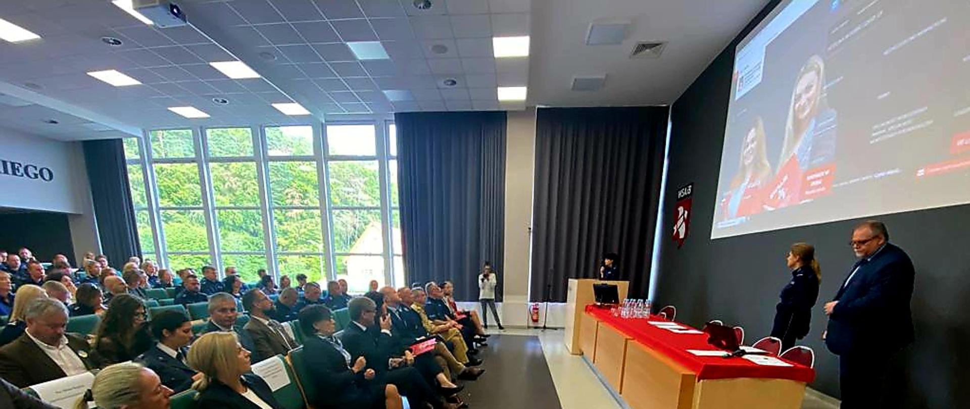 Funkcjonariusze Policji, Państwowej Straży Pożarnej oraz zaproszeni goście siedzą na auli oraz słuchają osób mówiących.