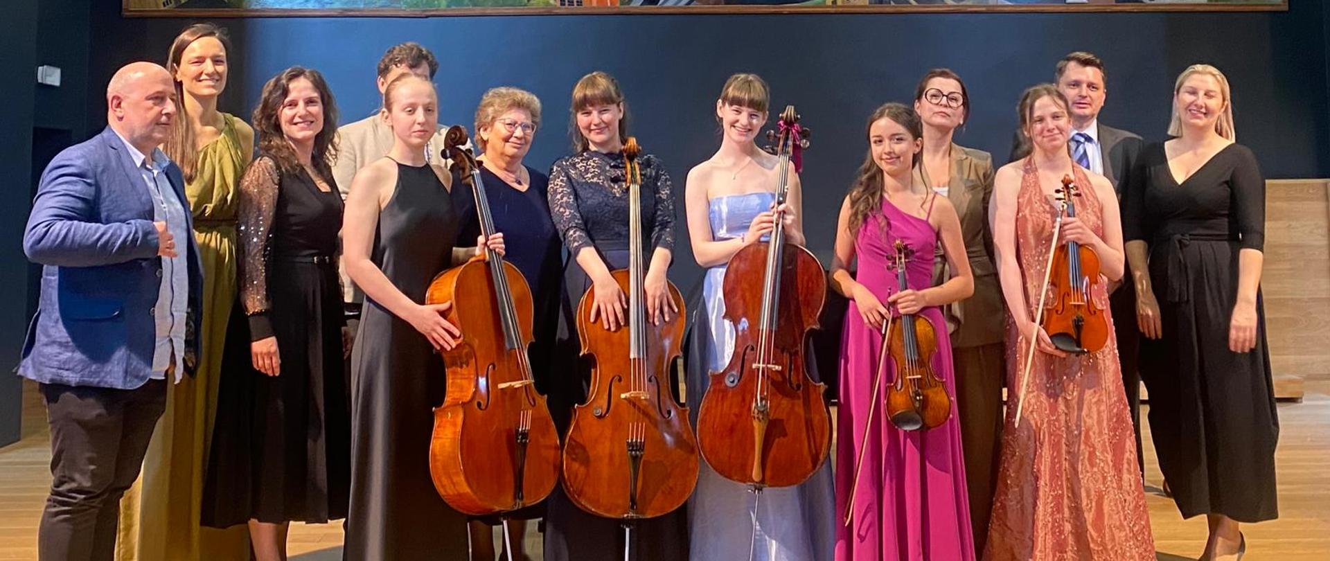 Kolorowe zdjęcie na scenie w dwóch nieregularnych rzędach stoją dyplomanci sekcji smyczkowej ich nauczyciele oraz pianistki - akompaniatorki. Wszyscy ubrani w galowe stroje z uśmiechami na twarzach. Dwie skrzypaczki trzymają w rękach skrzypce, a przed wiolonczelistkami stoją wiolonczele. W tle czarna ściana z kolorowym dużym obrazem przedstawiającym najważniejsze budynki i miejsca miasta Rybnika.