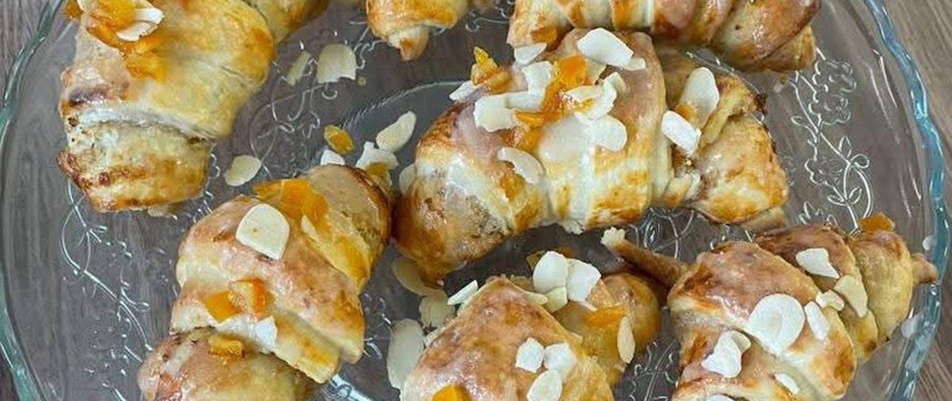 Na zdjęciu widać osiem małych, świeżo upieczonych rogalików, ułożonych na przezroczystym, szklanym talerzu z falowanym brzegiem. Talerz spoczywa na drewnianym stole o jasnobrązowym odcieniu.