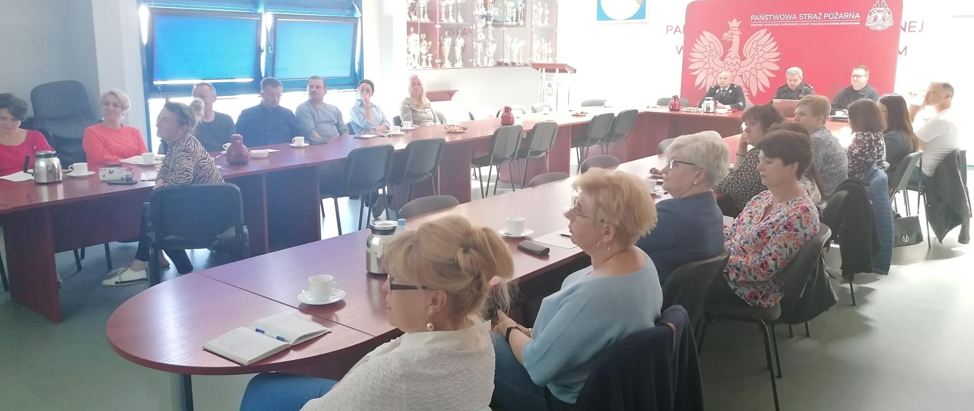 Uczestnicy spotkania siedzą przy stołach podczas narady kadry oświaty. Prezentacja na ścianie.