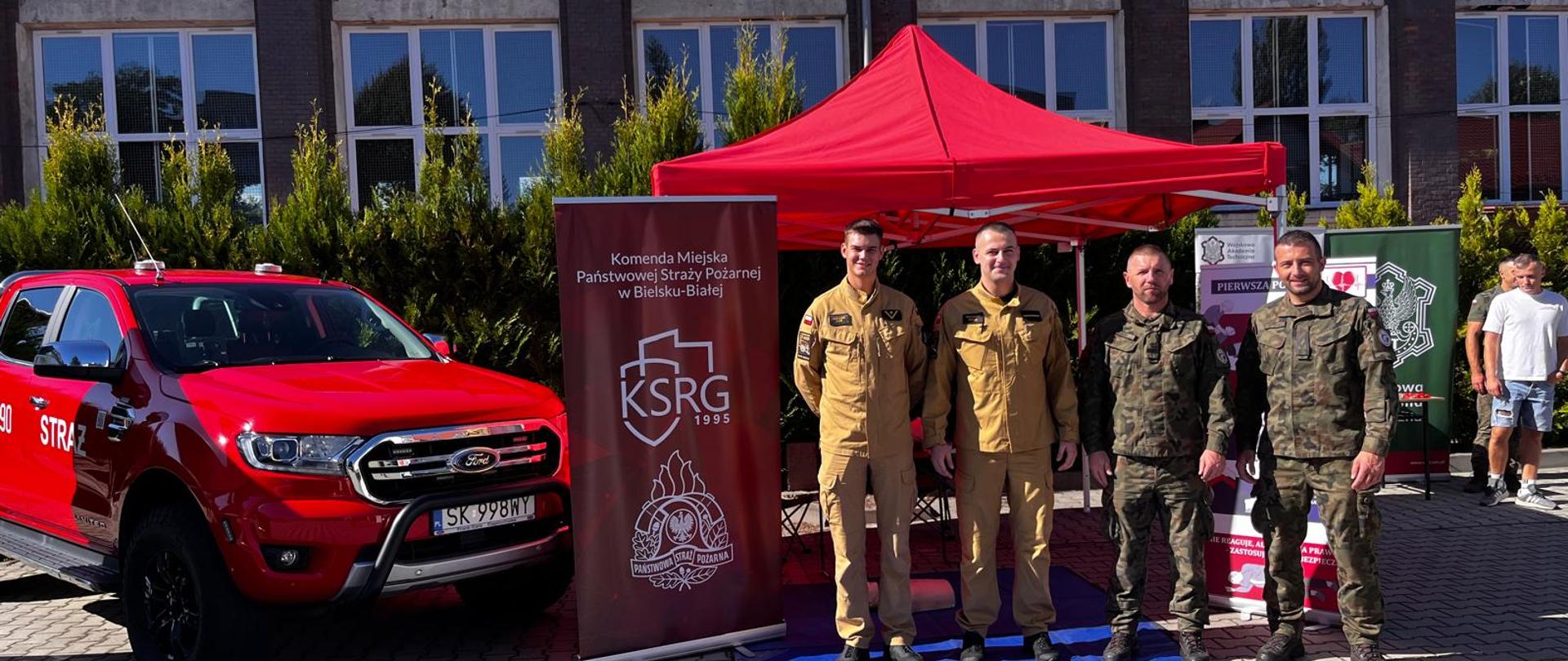 Opisane zdjęcie przedstawia czterech mężczyzn w mundurach wojskowych i strażackich, stojących przed budynkiem. Za nimi znajduje się czerwony namiot oraz czerwony pickup marki Ford, na którym widać napisy "STRAŻ". Obok stoją banery z logo Komendy Miejskiej Państwowej Straży Pożarnej w Bielsku-Białej oraz logo KSRG. 