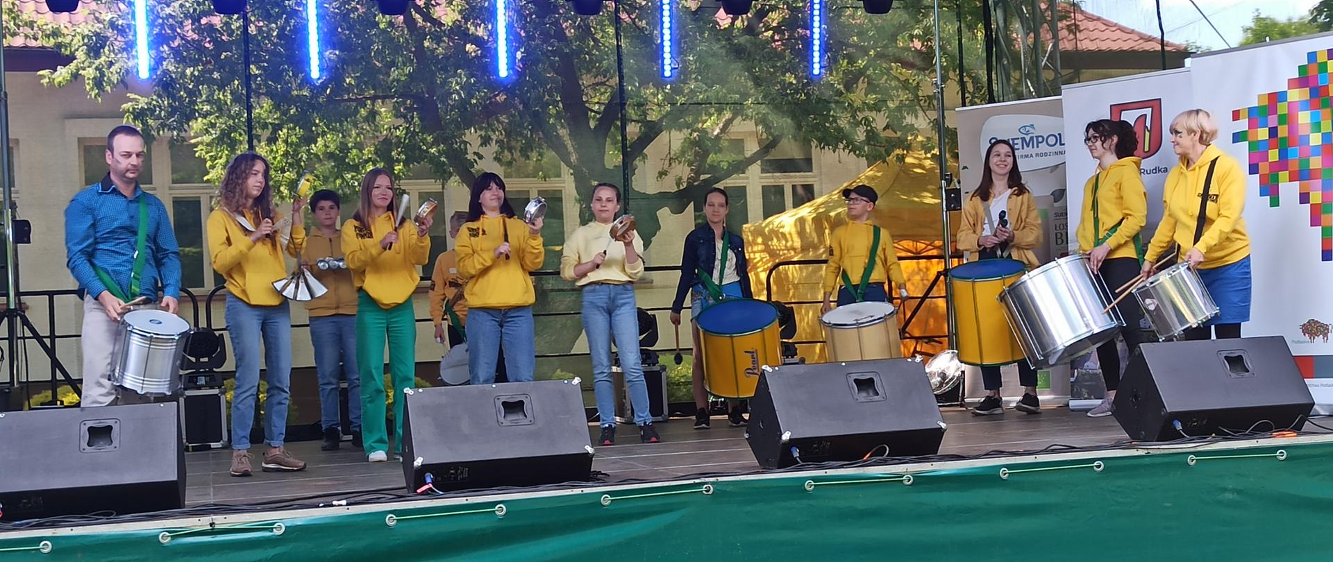 Uczniowie w żółtych bluzach stoją na scenie z różnymi instrumentami perkusyjnymi na których grają. Prowadzący Batucadę w niebieskiej koszuli z bębnem zawieszonym na pasie przez ramię, stoi po lewej stronie.