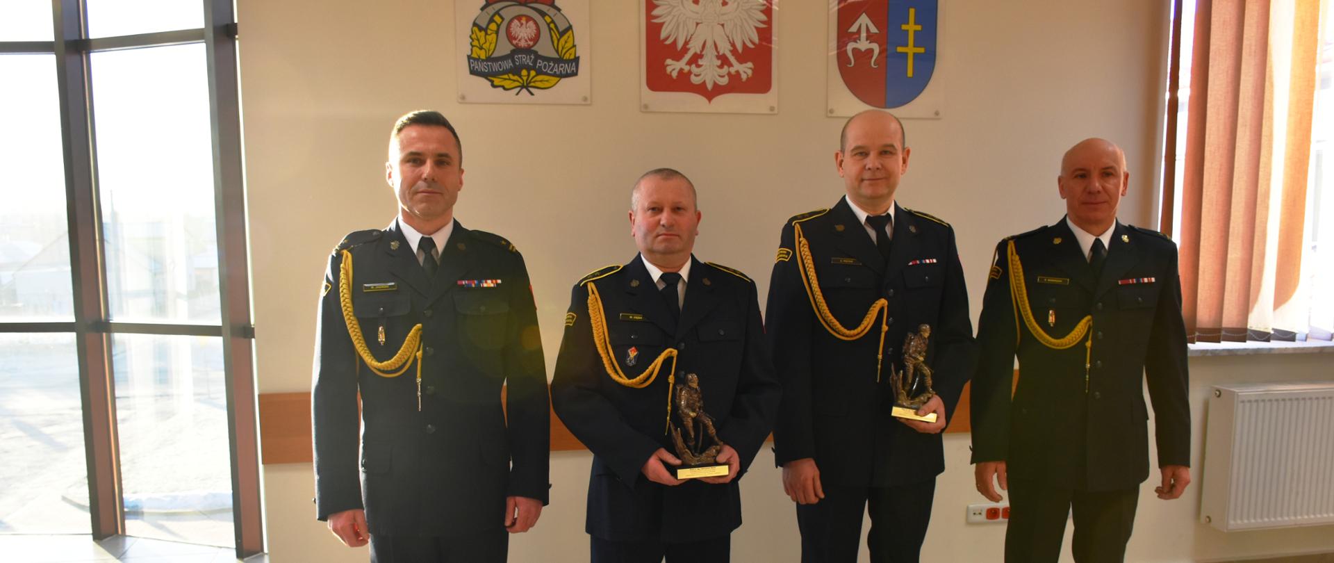 Zdjęcie dwóch strażaków odchodzących na zaopatrzenie emerytalne. W rękach trzymają pamiątkowe statuetki, a po ich prawej i lewej stronie stoi komendant oraz z-ca komendanta powiatowego. Zdjęcie wykonane na auli.