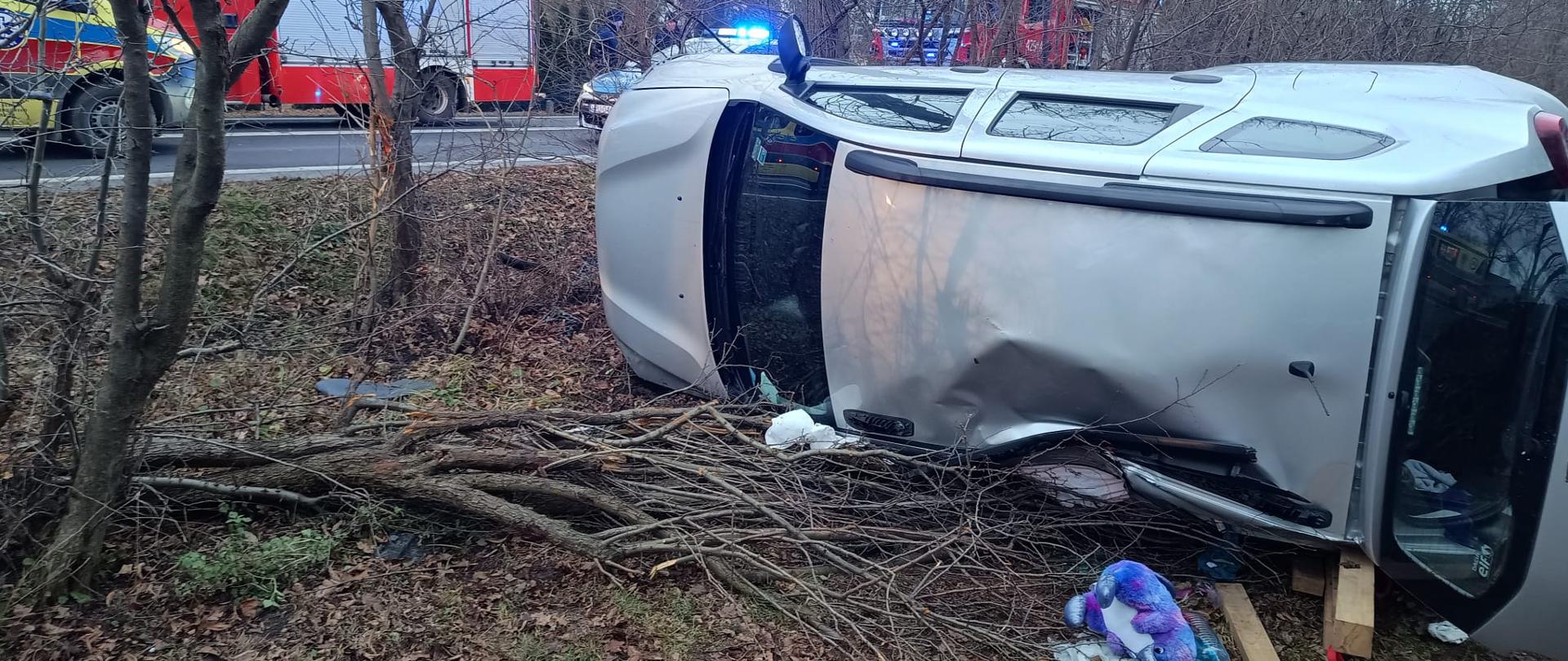 Na zdjęciu widać dach uszkodzonego pojazdu, który znajduje się poza drogą w pozycji na lewym boku. W tle na drodze stoją samochody służb ratowniczych