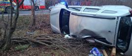 Na zdjęciu widać dach uszkodzonego pojazdu, który znajduje się poza drogą w pozycji na lewym boku. W tle na drodze stoją samochody służb ratowniczych