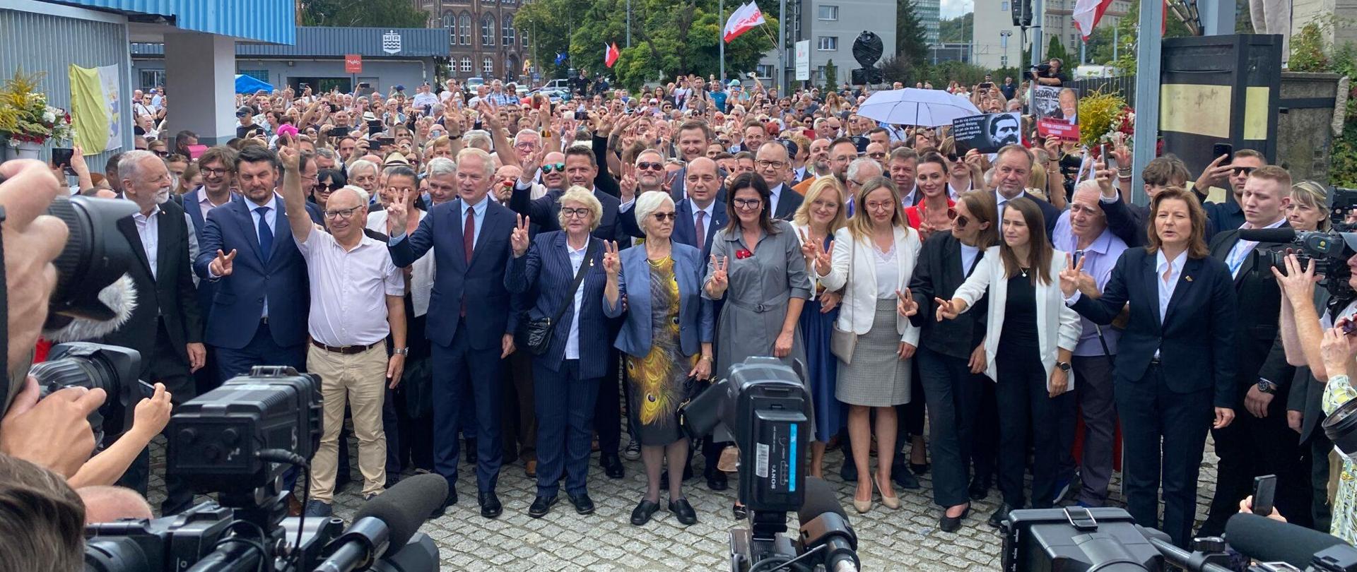 Tłum ludzi przed bramą stoczni, w tle biało czerwone flagi