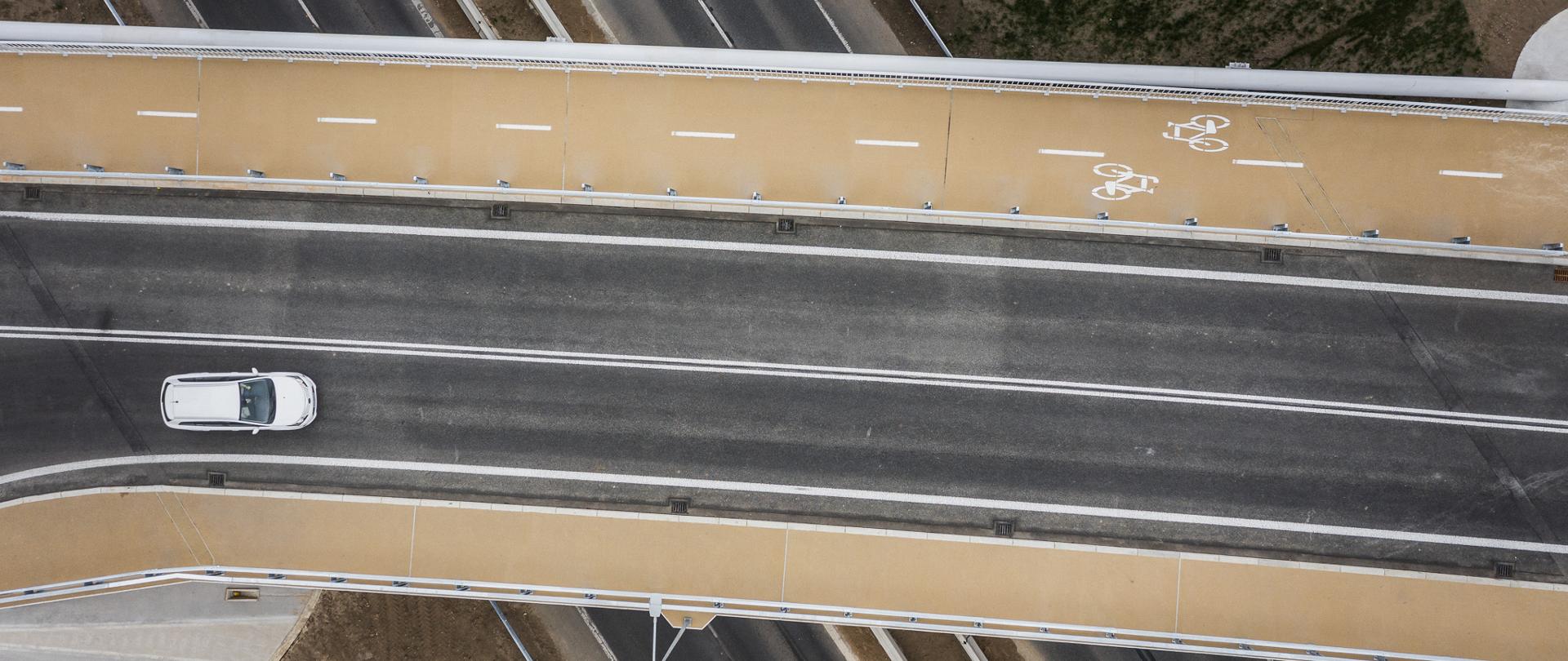 Widok z góry na wiadukt nad DK86 z wyznaczonym ciągiem pieszo-rowerowym; pod obiektem widoczne jezdnie drogi krajowej i przejeżdżające pojazdy.