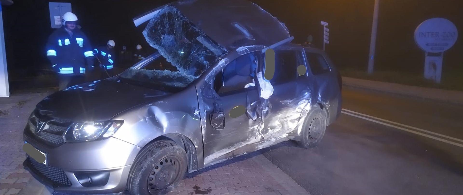 Na zdjęciu wykonanym w porze nocnej widzimy samochód osobowy postawiony na koła, stojący w poprzek drogi. Posiada on zniszczony bok oraz częściowo odcięty dach przez który strażacy prowadzili ewakuację poszkodowanych. W tle widoczni strażacy usuwają rozbite części pojazdu z jezdni.