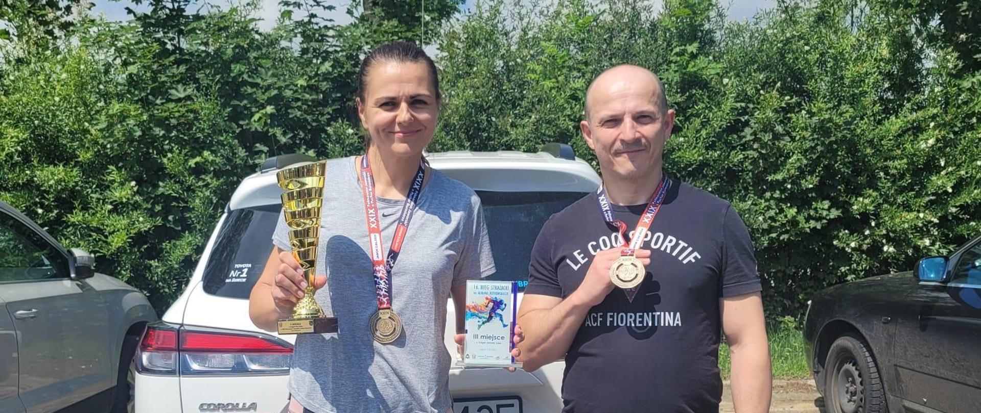 Kobieta i mężczyzna stoją na tle auta. Kobieta w ręku trzyma puchar a mężczyzna medal.
