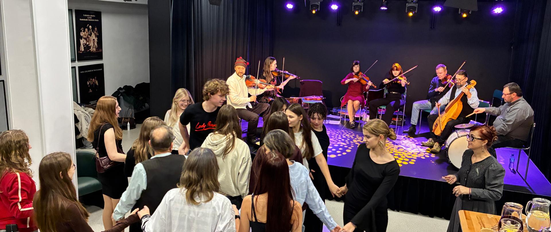 W dużym pomieszczeniu na scenie siedzi siedmioro dorosłych muzyków, pięcioro gra na skrzypcach, jeden na kontrabasie i jeden na bębnie (siedzi z prawej strony). Przed sceną grupa kilkunastu młodych ludzi tańczy trzymając się za ręce. W środku widać również dorosłego mężczyznę i kobietę stojącą z boku po prawej stronie. Na stoliku z prawej strony stoją dzbanki i naczynia z napojami. Na górze widać zainstalowane duże reflektory oświetleniowe na szynach.