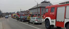 Kolorowe zdjęcie wykonane w pochmurny dzień przedstawia drogę, na której stoją częściowo widoczne czerwone pojazdy straży pożarnej i karetka pogotowia ratunkowego. Pomiędzy pojazdami służb ratowniczych stoją uszkodzone pojazdy osobowe. W tle widać szary budynek remizy strażackiej z wysokim masztem antenowym na dachu.