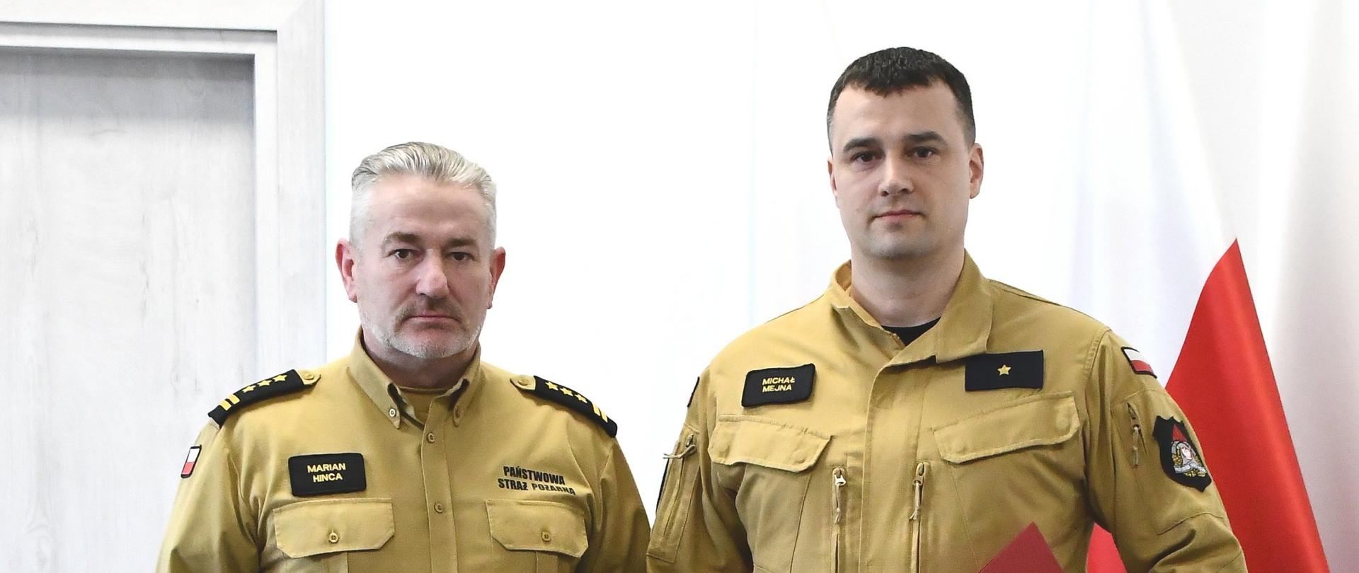 Strażacy w mundurach koloru musztardowego stoją obok siebie jeden z nich trzyma czerwoną teczką za nimi ustawione są dwie flagi Polski.