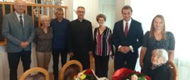 Na fotografii widać grupę stojących osób. W centrum, przy stole z bukietami kwiatów siedzi uśmiechnięta starsza kobieta – jubilatka obchodząca setne urodziny. Obok niej stoją członkowie rodziny oraz przedstawiciele lokalnych władz i instytucji.