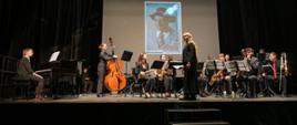 Cały skład big bandu na scenie Teatru Witkacy. Dyrygent stoi bokiem do widowni przed nią uczniowie siedzą w dwóch rzędach ze swoimi instrumentami, przed nimi pulpity, na której ustawione są nuty. Na ścianie w tle zdjęcie Stanisława Witkacego.