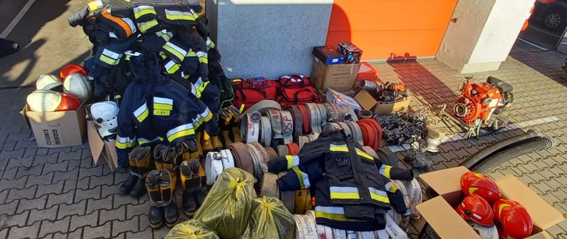 Fotografia ukazuje ujęcie placu przed garażami Jednostki Ratowniczo-Gaśniczej w Strzelcach Krajeńskich. Na placu złożono sprzęt do przekazania ukraińskim strażakom. Wśród sprzętu znajdują się hełmy, ubrania specjalne, węże pożarnicze, prądownice, i inne.