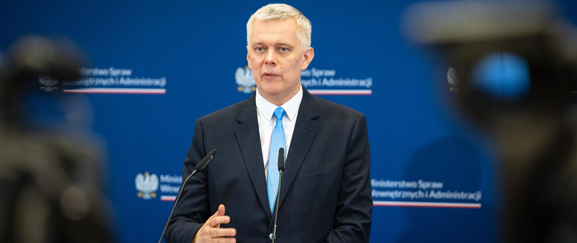 Minister Tomasz Siemoniak stoi na tle niebieskiej ścianki ze zmuliplikowanym logo MSWiA (orzeł, napis Ministerstwo Spraw Wewnętrznych i Administracji oraz biało-czerwony pasek). Przed ministrem dwa mikrofony kierunkowe. Po prawej i lewej stronie zdjęcia widać rozmazane kamery telewizyjne. 