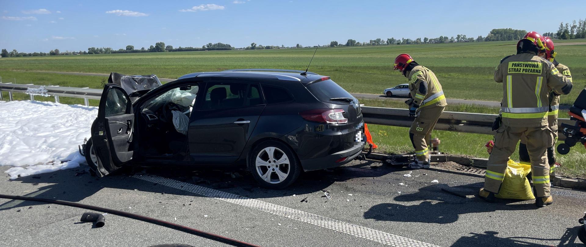 uszkodzony czarny samochód na poboczu drogi za nim strażacy