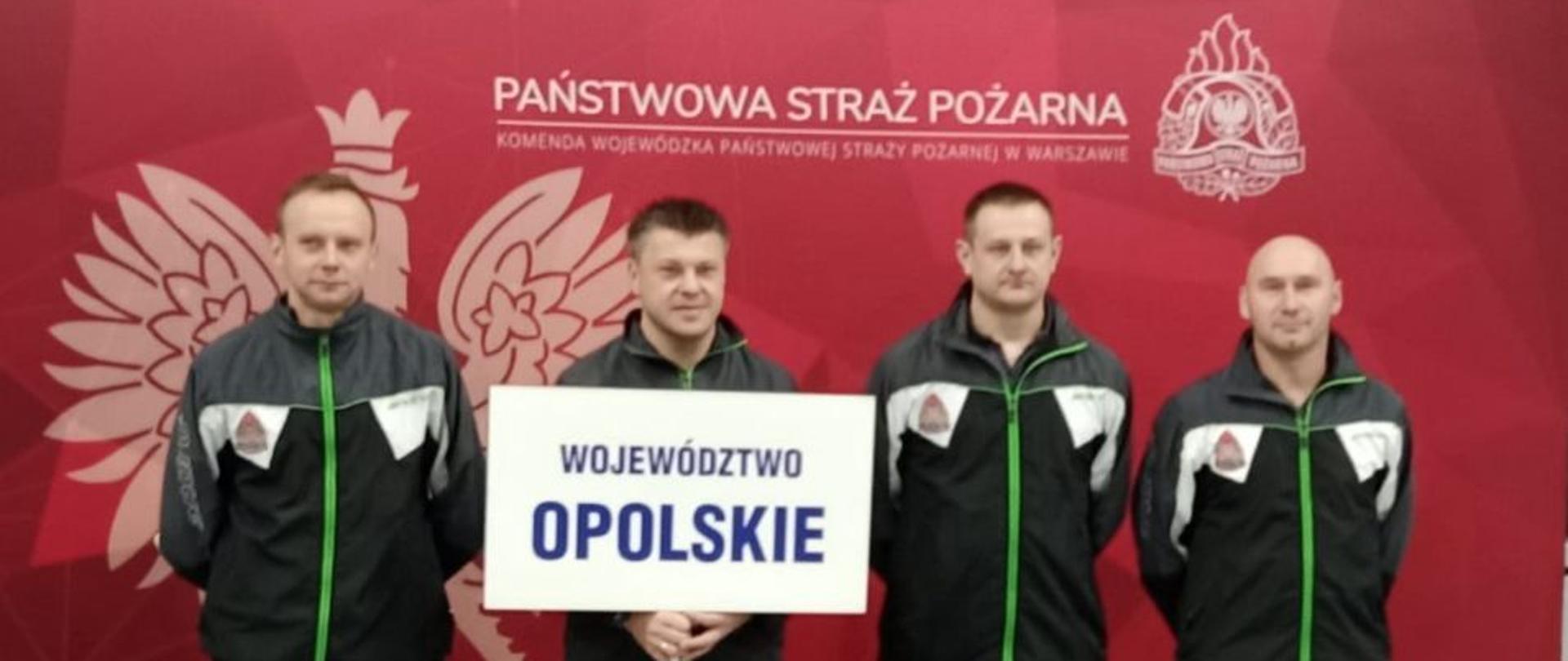 Reprezentacja Państwowej Straży Pożarnej województwa opolskiego. Strażacy w strojach sportowych stoją na tle banneru Państwowej Straży Pożarnej koloru czerwonego trzymając tablicę białą z napisem: województwo opolskiego.