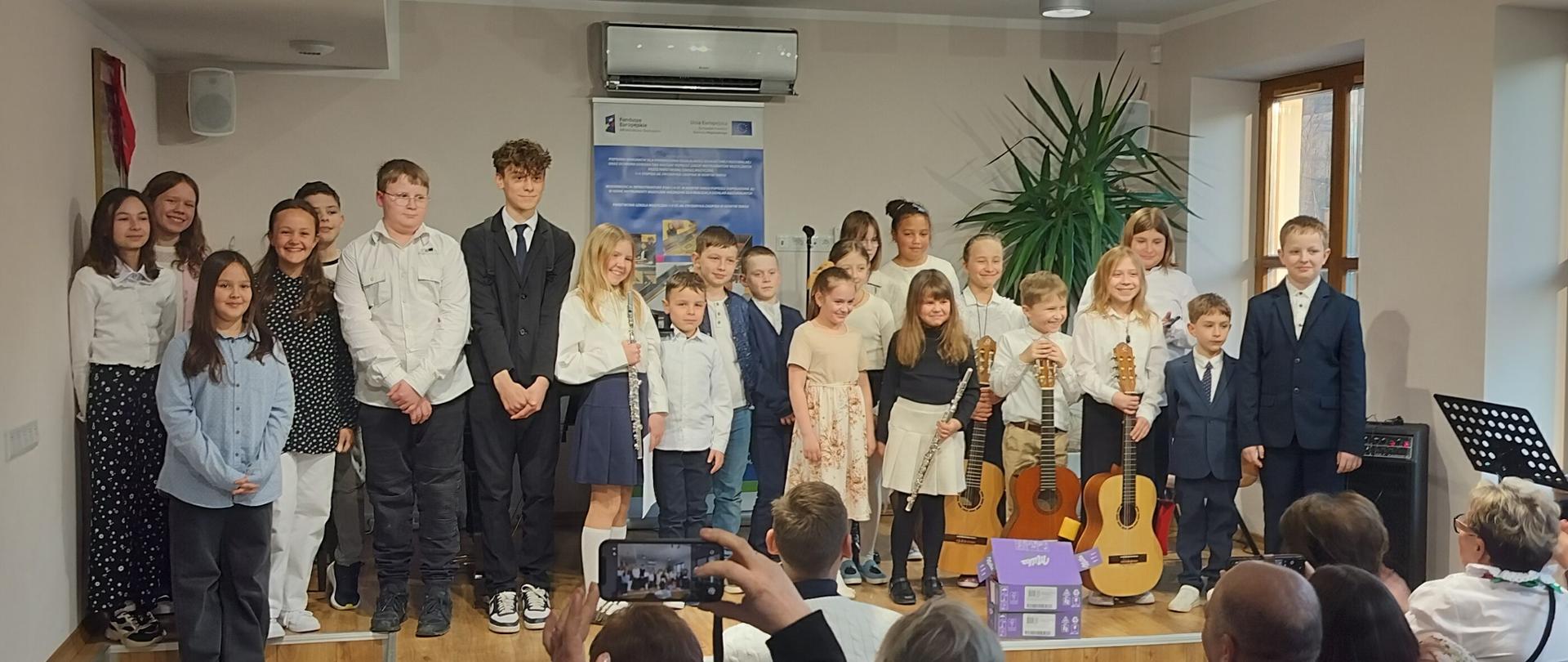 Zdjęcie przedstawia grupę dzieci i młodzieży stojących na niskim podestcie, prawdopodobnie podczas występu lub uroczystości w kameralnej sali. Uczestnicy: Na scenie znajduje się około dwudziestu młodych osób w różnym wieku, ubranych przeważnie w stroje galowe (białe koszule, ciemne spodnie, spódnice, a niektórzy w garnitury). Instrumenty: Część dzieci trzyma instrumenty muzyczne – widoczne są gitary klasyczne oraz flety poprzeczne, co sugeruje, że jest to koncert lub zakończenie warsztatów muzycznych. Otoczenie: W tle widać niebieski baner z informacjami (prawdopodobnie o projekcie unijnym), dużą roślinę doniczkową oraz klimatyzator. Na dole zdjęcia widoczne są głowy i ręce publiczności robiącej zdjęcia telefonami.Atmosfera: Wydarzenie ma charakter oficjalny, ale radosny; dzieci wyglądają na dumne ze swojego występu.