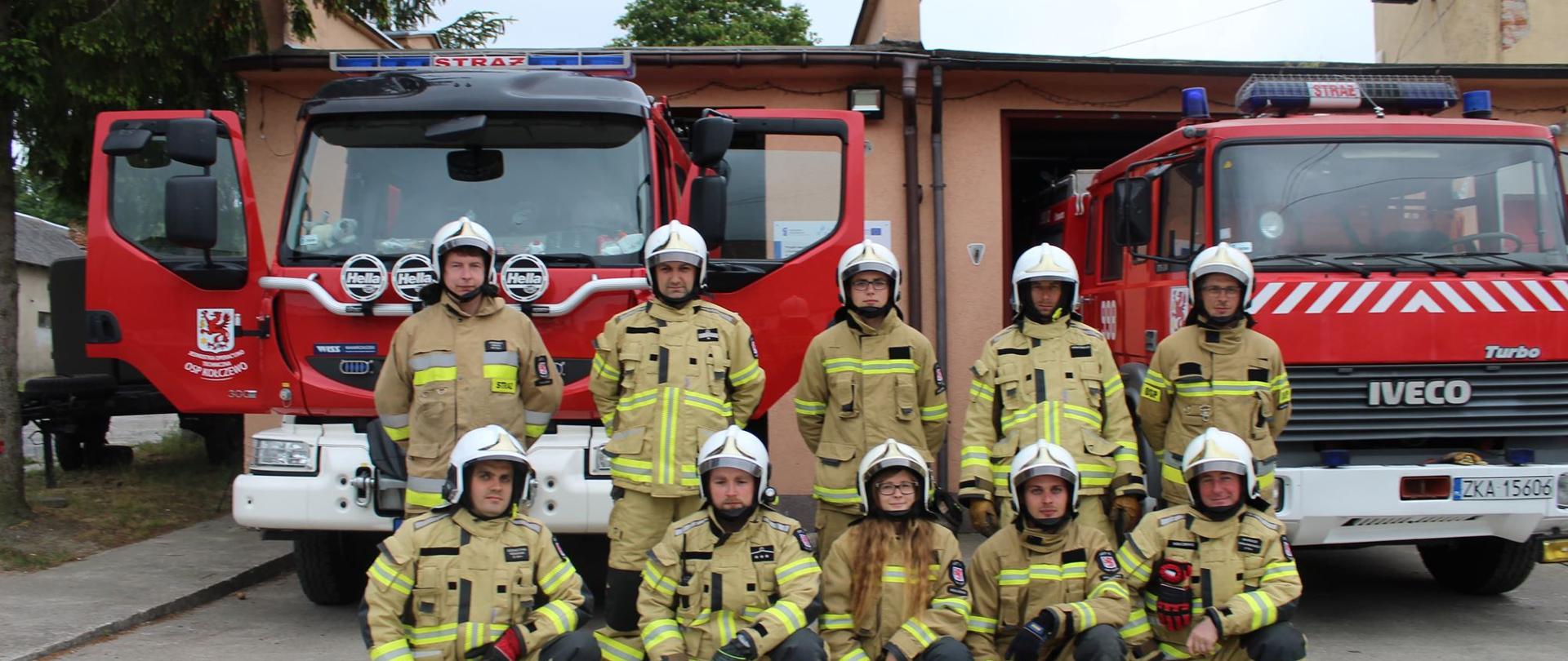 Zdjęcie przedstawia strażaków OSP Kołczewo w ubraniach specjalnych na tle wozów strażackich i remizy.